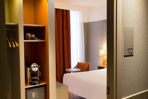 A hotel room with a bed, a coffee machine, a small table, and a wardrobe with hanging items.