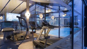 Un gimnasio moderno con equipamiento de ejercicio, una pantalla grande y vistas a una piscina a través de paredes de vidrio.