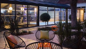 An outdoor seating area with chairs and a table, a glass building with interior lighting, and a small tree. - Hôtel Oceania Le Jura Dijon (Dijon)