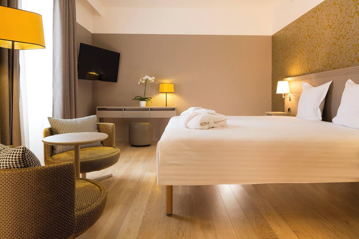 A hotel room with a large bed, two bedside tables with lamps, a flat-screen TV mounted on the wall, a small round table with a chair, and a potted plant.