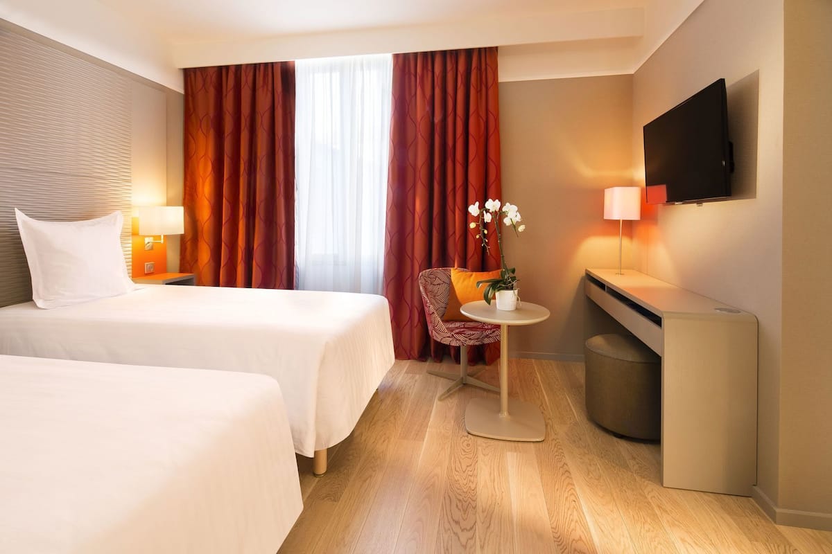 A hotel room with two beds, a desk, a chair, a TV, and a window with curtains.