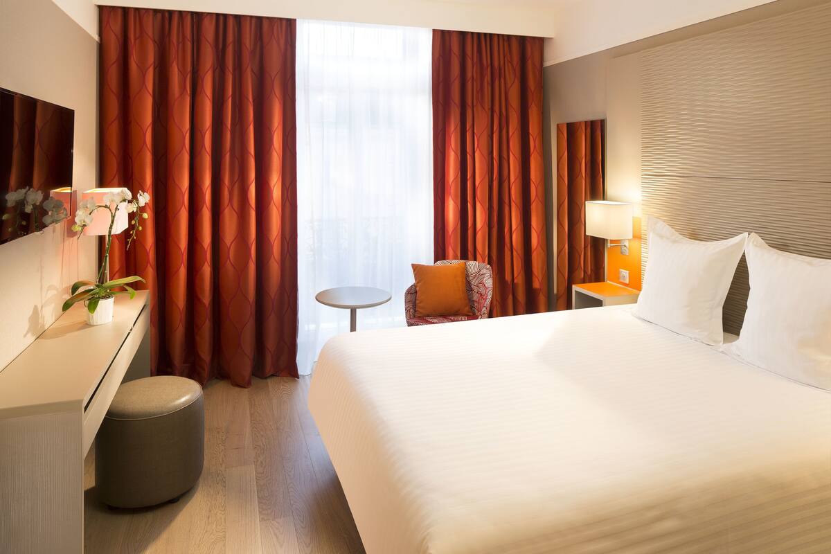 A hotel room with a large bed, a desk, a chair, a small table, and a lamp.