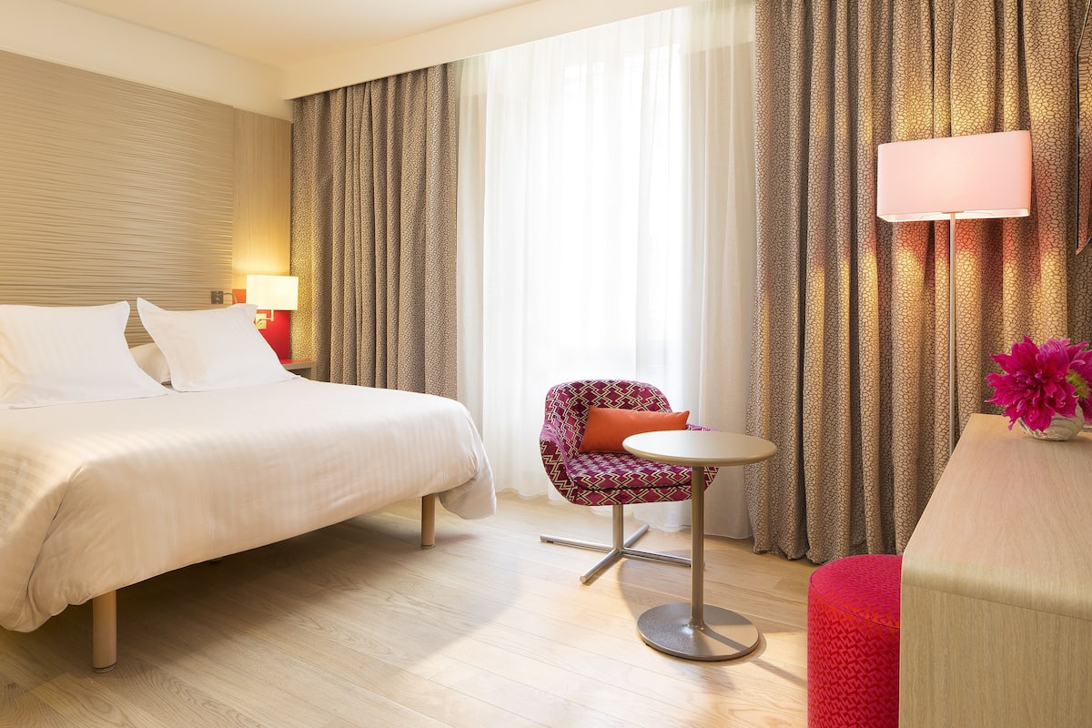 A hotel room with a bed, a small round table, a chair, and a desk with a lamp.