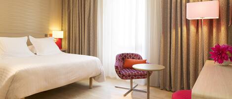 A hotel room with a bed, a small round table, a chair, and a desk with a lamp.