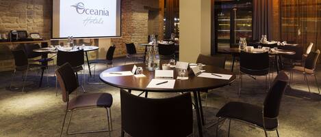 A conference room with a presentation screen displaying 'Oceania Hotels' logo, round tables with chairs, and a brick wall.