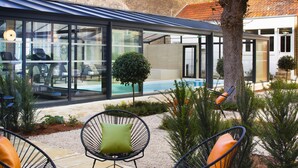 A modern outdoor seating area with chairs and a table, a swimming pool, and a glass-enclosed building. - Hôtel Oceania Le Jura Dijon (Dijon)