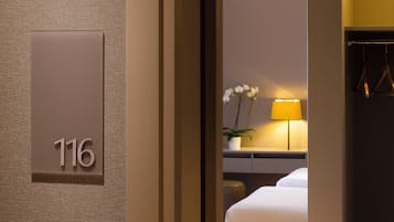 Une chambre d’hôtel avec le numéro « 116 » affiché sur le mur, un lit, une table de chevet avec une lampe et une plante en pot.