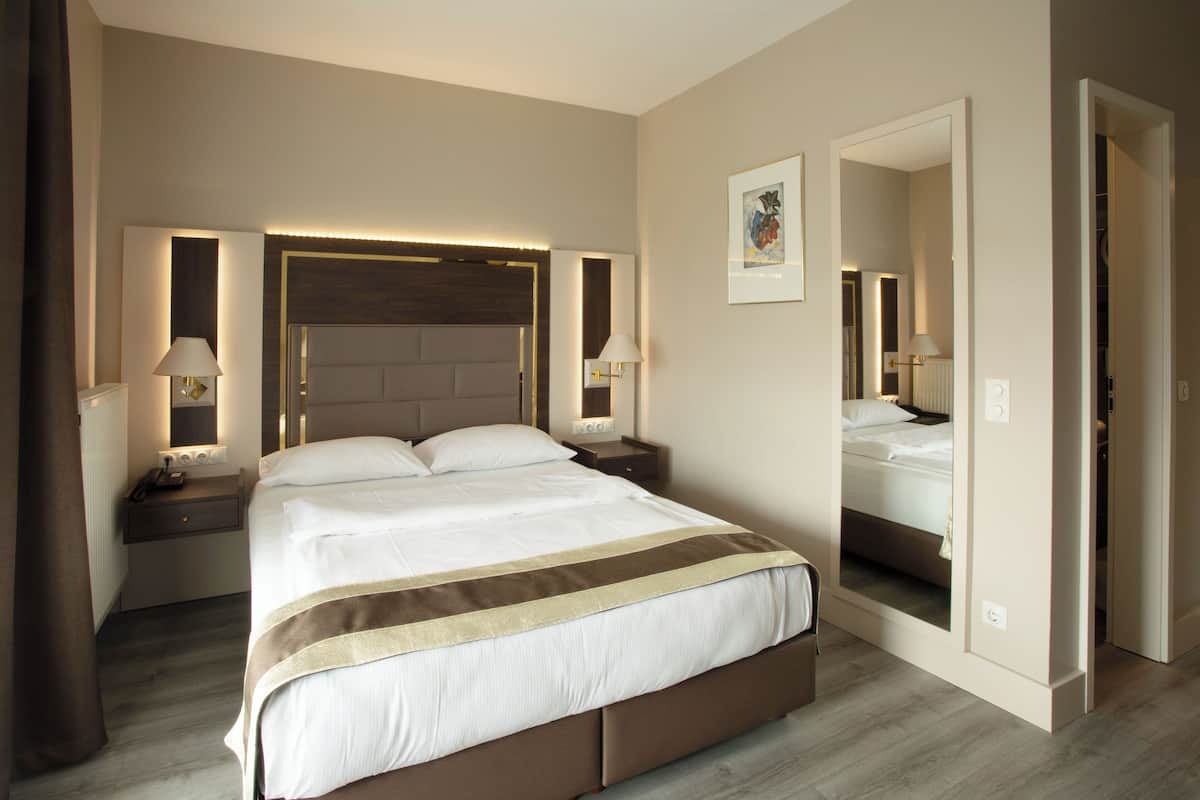 Une chambre d’hôtel avec un grand lit, des tables de chevet avec des lampes, une tête de lit dotée d’un éclairage intégré, un miroir et une porte donnant sur une autre pièce.
