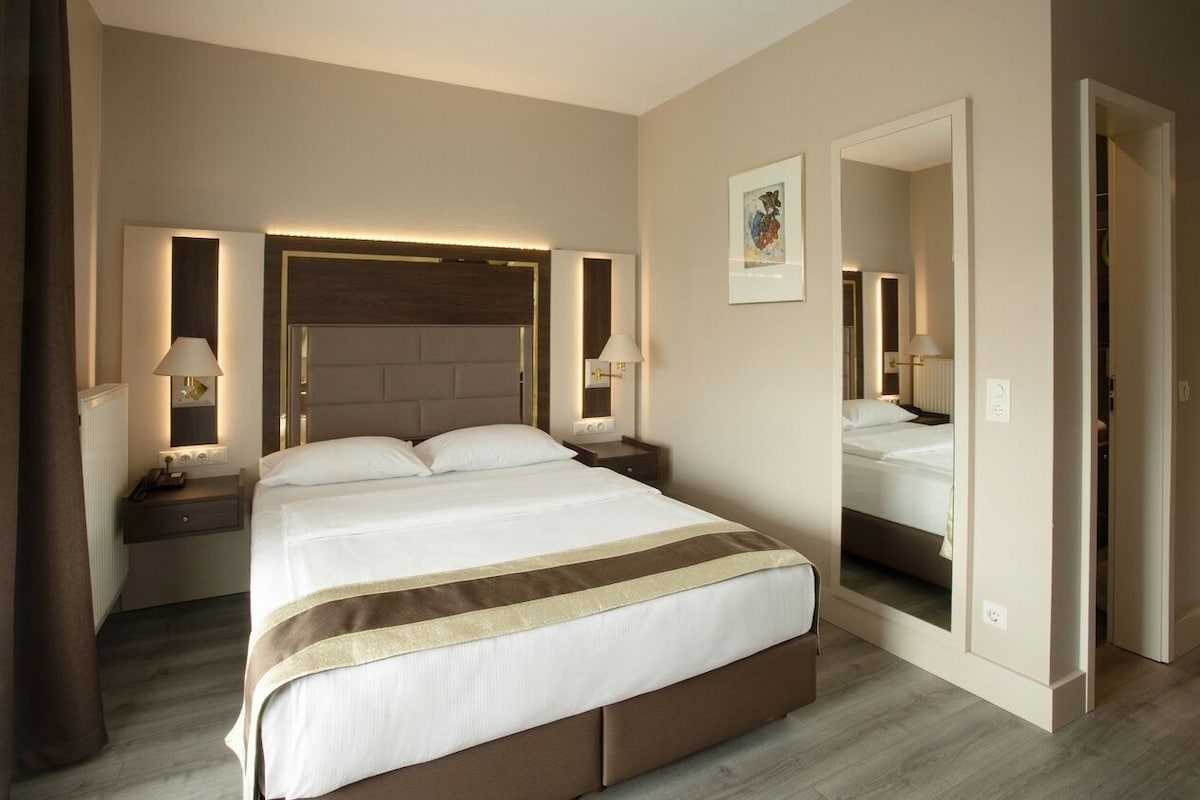 Quarto de hotel com uma cama grande, mesas de cabeceira, um espelho e uma porta para um armário.