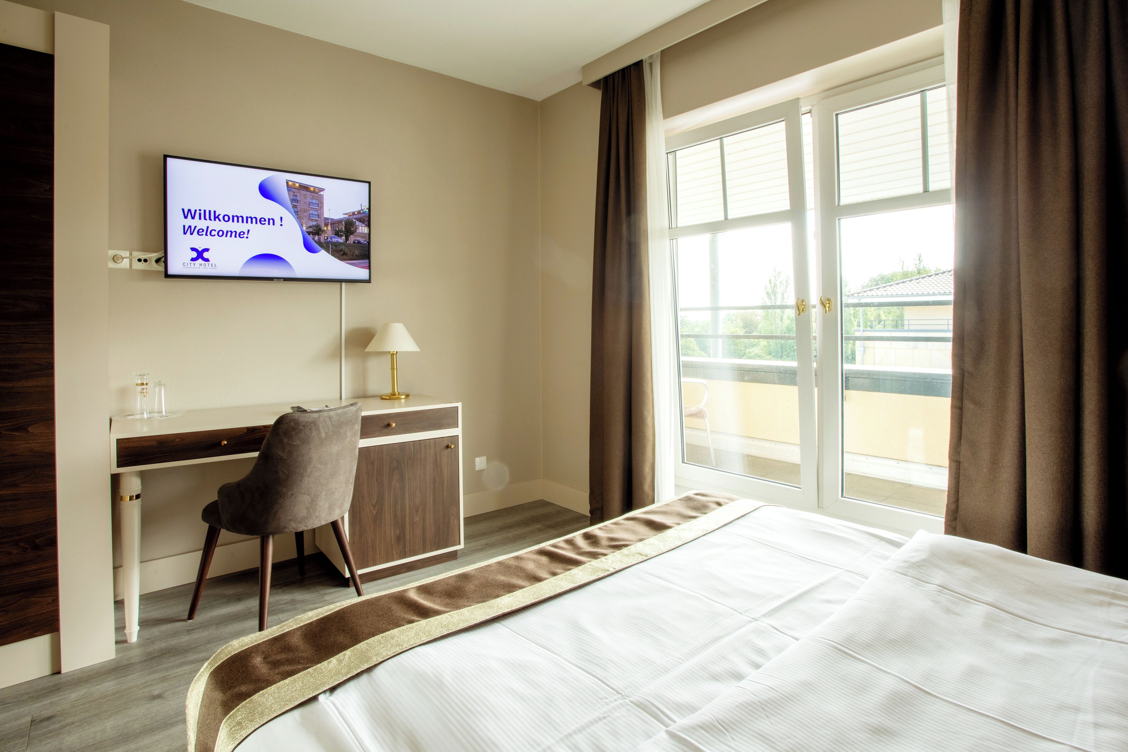 Habitación de hotel con cama, escritorio, silla, televisor mostrando un mensaje de bienvenida y ventana con cortinas.