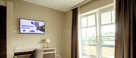 Habitación de hotel con cama, escritorio, silla, televisor mostrando un mensaje de bienvenida y ventana con cortinas.