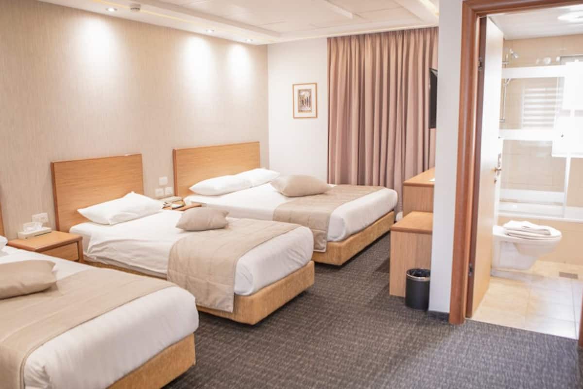 A hotel room with three single beds, a wooden headboard, a small bedside table with a lamp, a trash bin, and a view of the bathroom through a door.