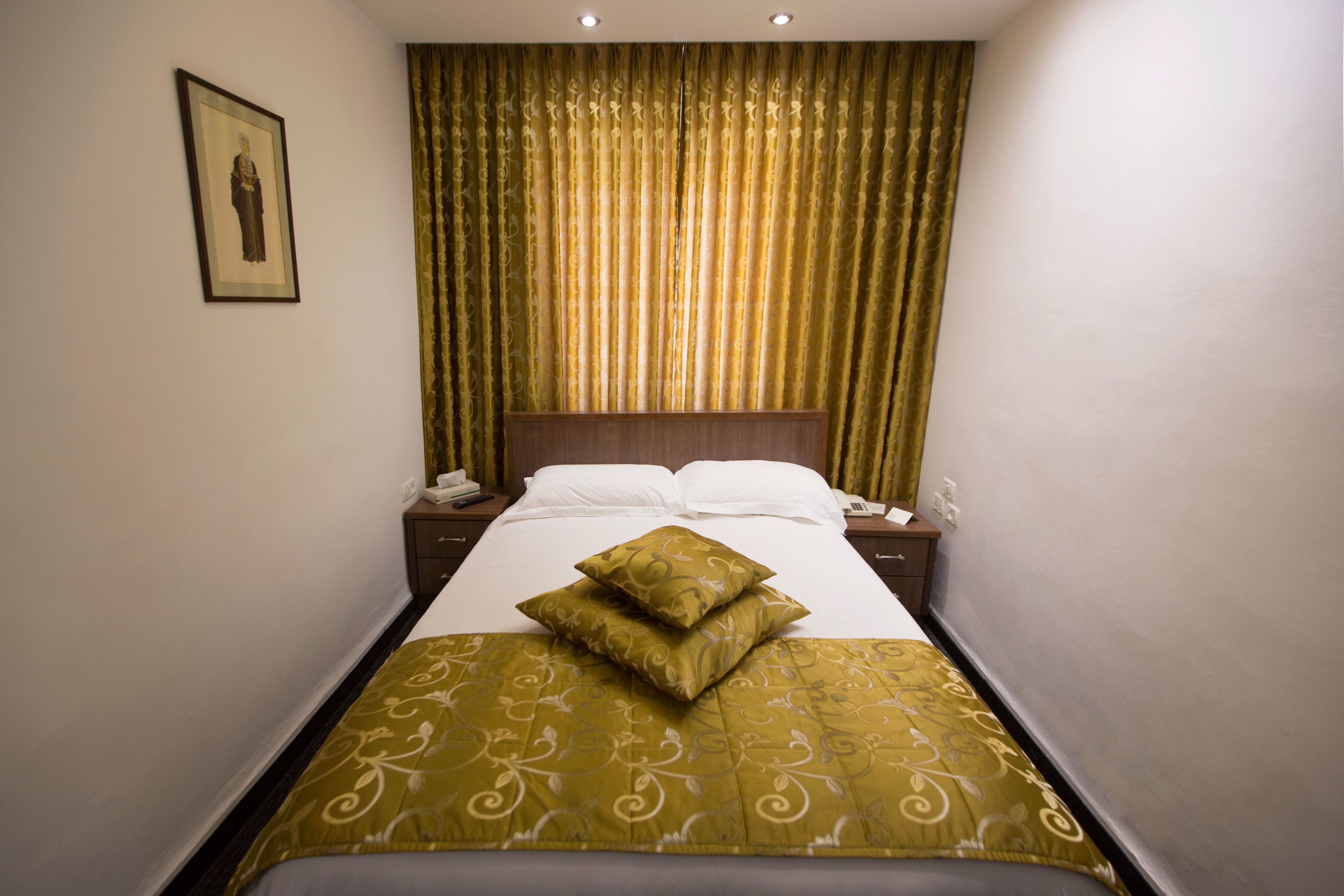 A narrow hotel room with a bed, a nightstand, a telephone, and a framed picture on the wall.