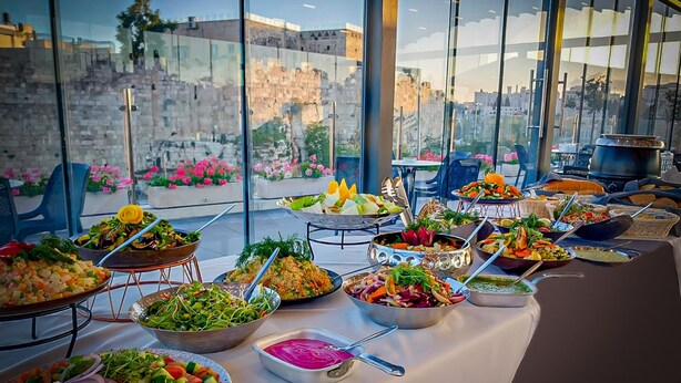 A buffet table with various dishes and a view of a historic stone wall and cityscape through large windows. - The Golden Walls Hotel (Jerusalem)