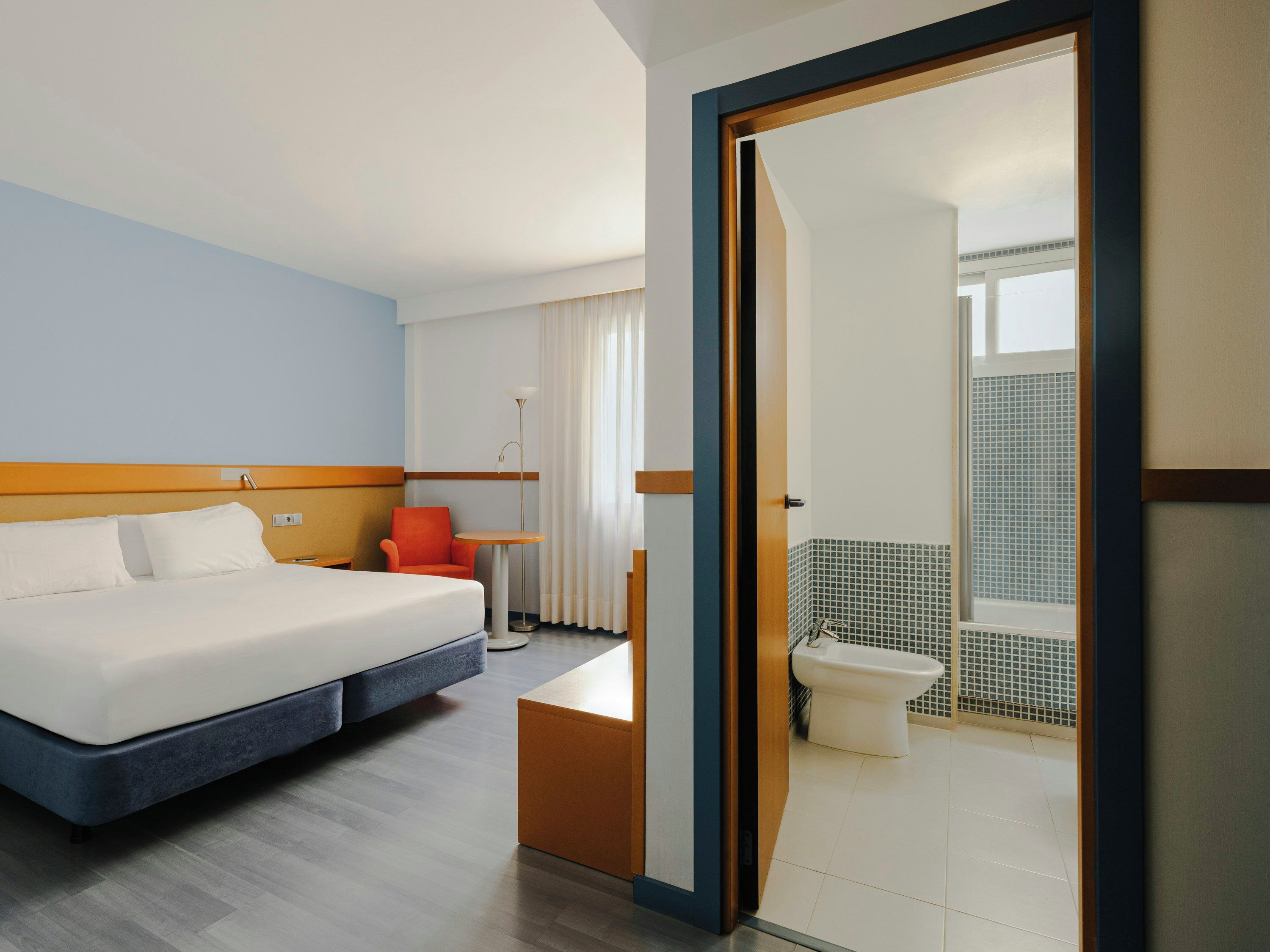 A hotel room with a bed, a chair, a small table, and a view of the bathroom.