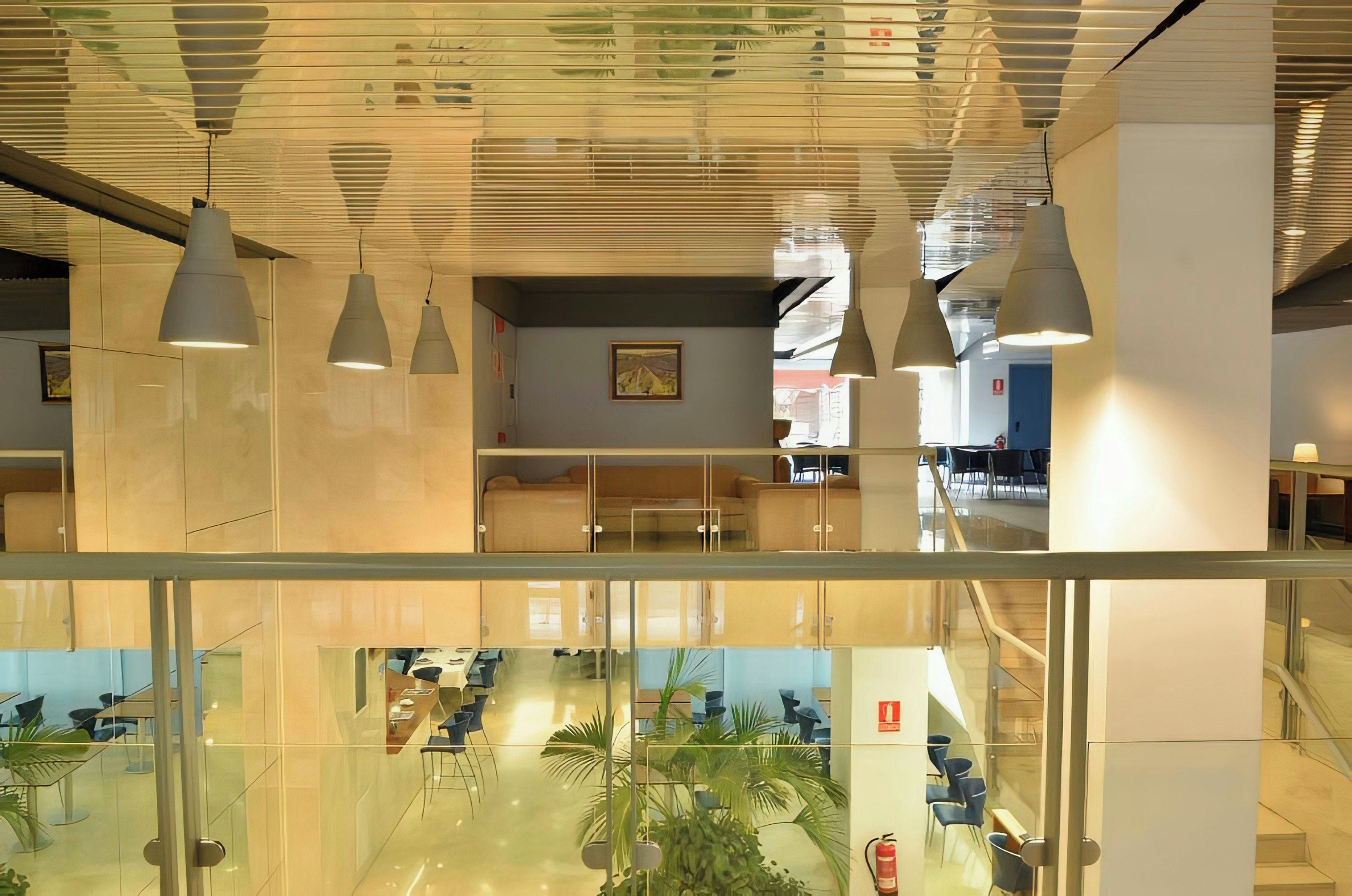 Un interior moderno con divisiones de vidrio, iluminación colgante y una zona de estar visible con mesas y sillas.