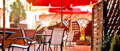 Un espace extérieur aménagé en terrasse, avec des chaises et des tables en métal, des parasols rouges et un mur en briques.