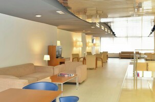Um saguão moderno de hotel com área de estar, balcão de recepção e uma grande janela que proporciona luz natural.