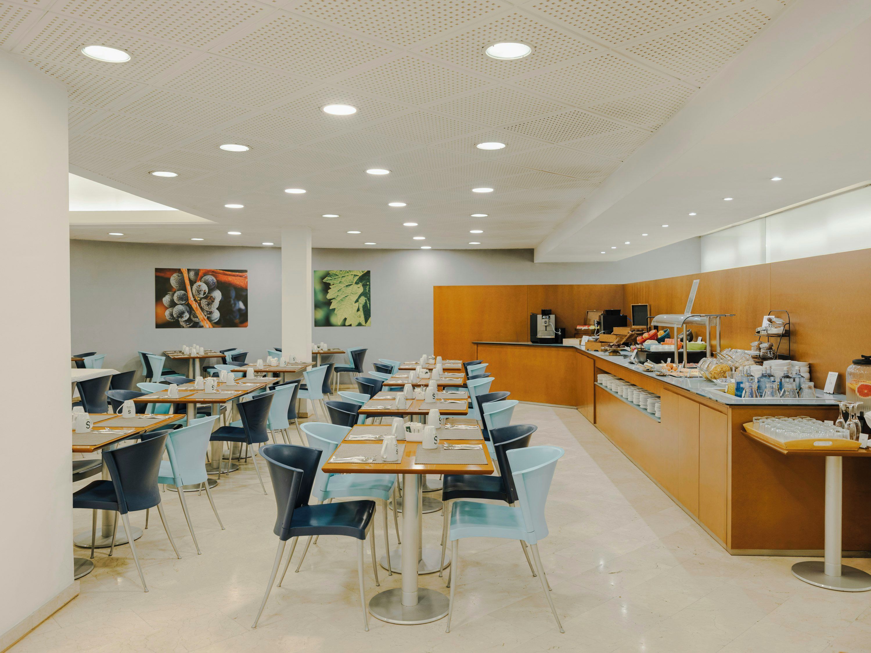 A modern cafeteria with multiple tables and chairs, a counter with food and beverages, and a ceiling with recessed lighting.