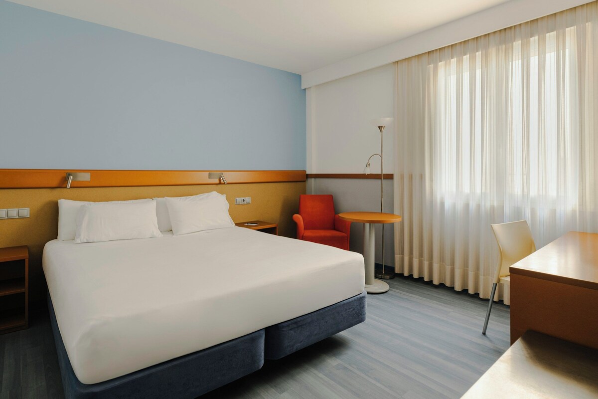 A hotel room with a large bed, a desk, a chair, a small table, and a floor lamp.