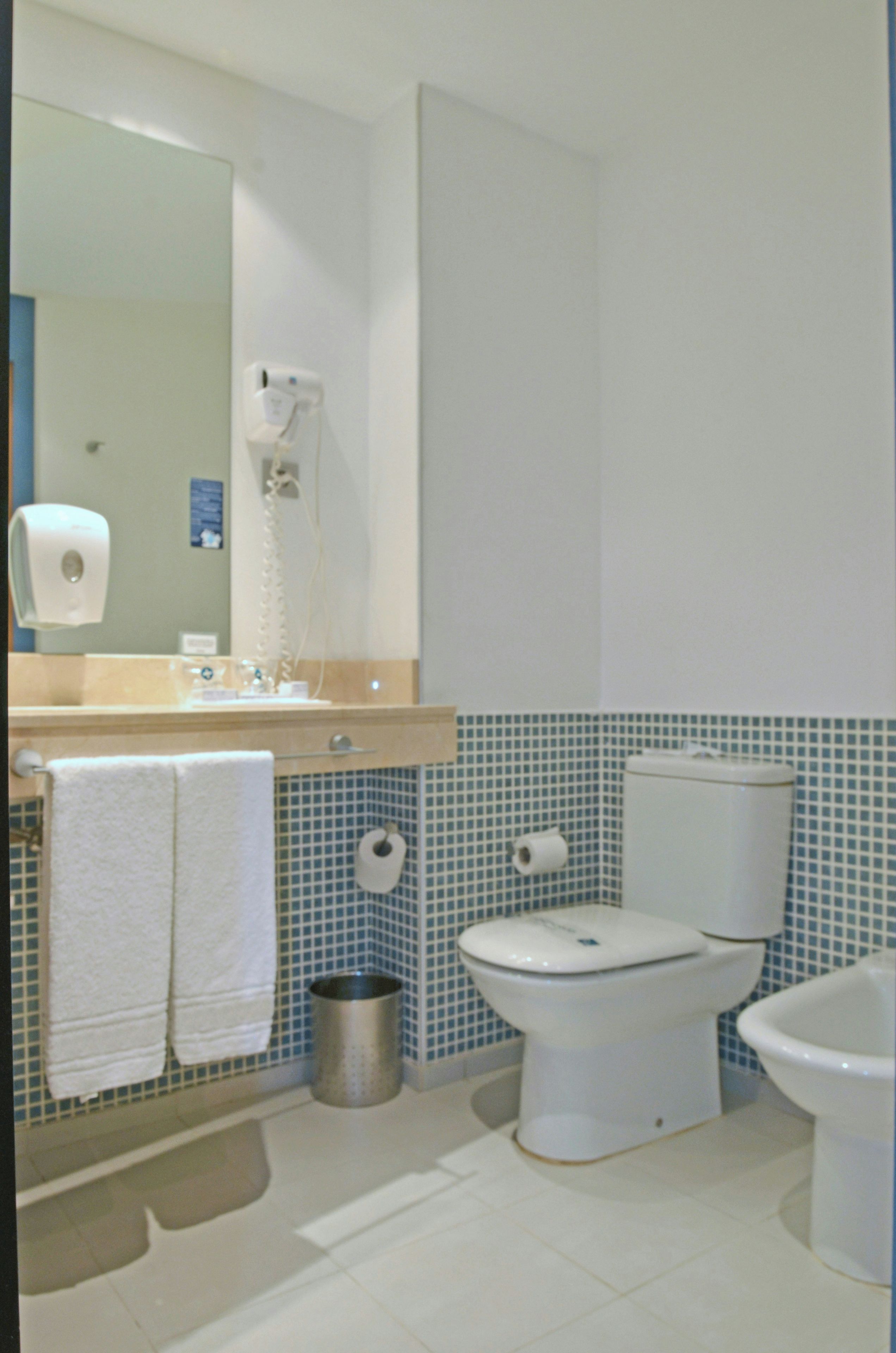 A bathroom with a toilet, bidet, and a hand dryer. There is a trash can and a towel rack.
