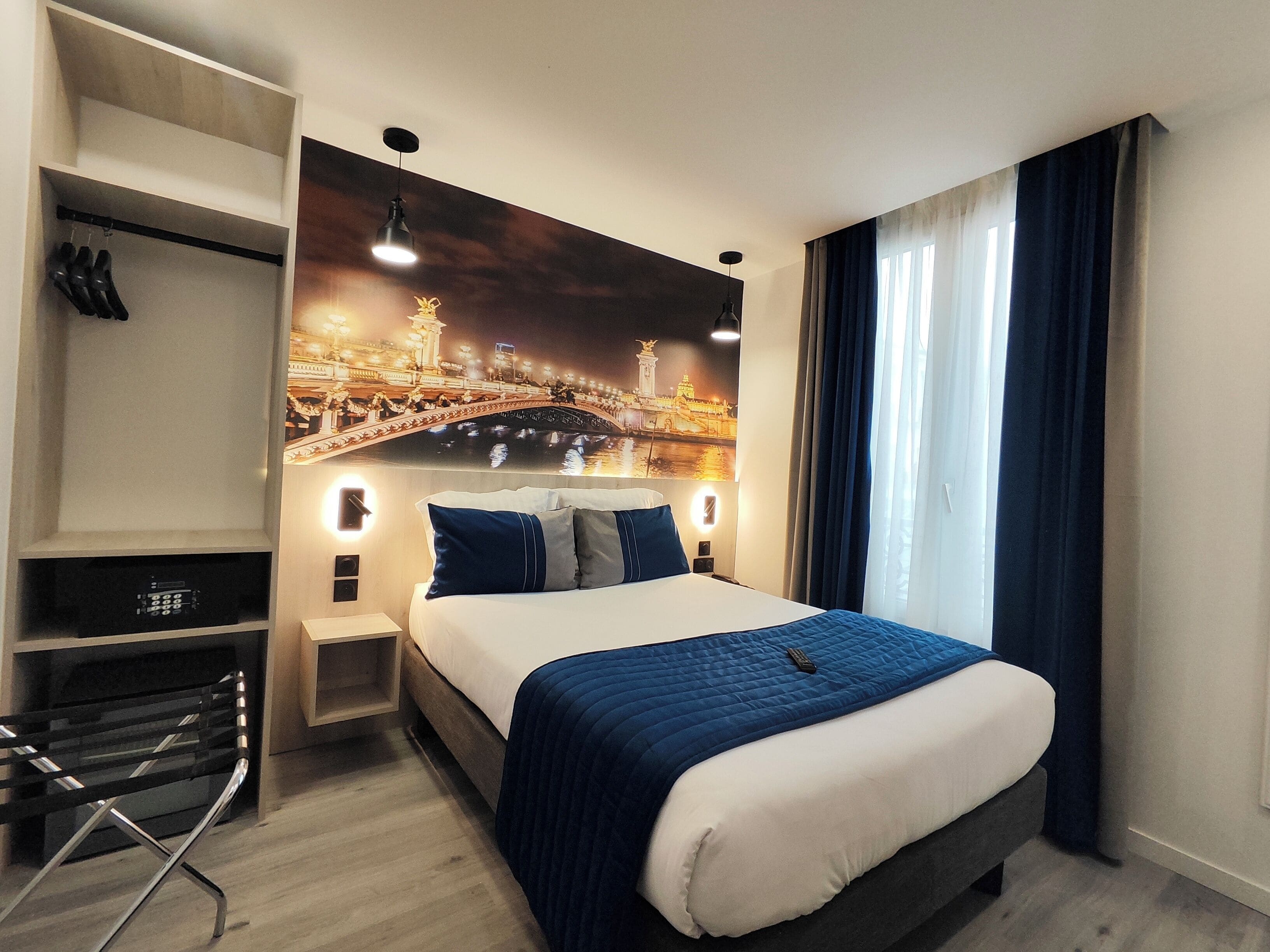 Ein Hotelzimmer mit einem Bett, einem Schreibtisch und einem Stuhl. Eine Wandhalterung zeigt Schuhe und eine Fernbedienung.