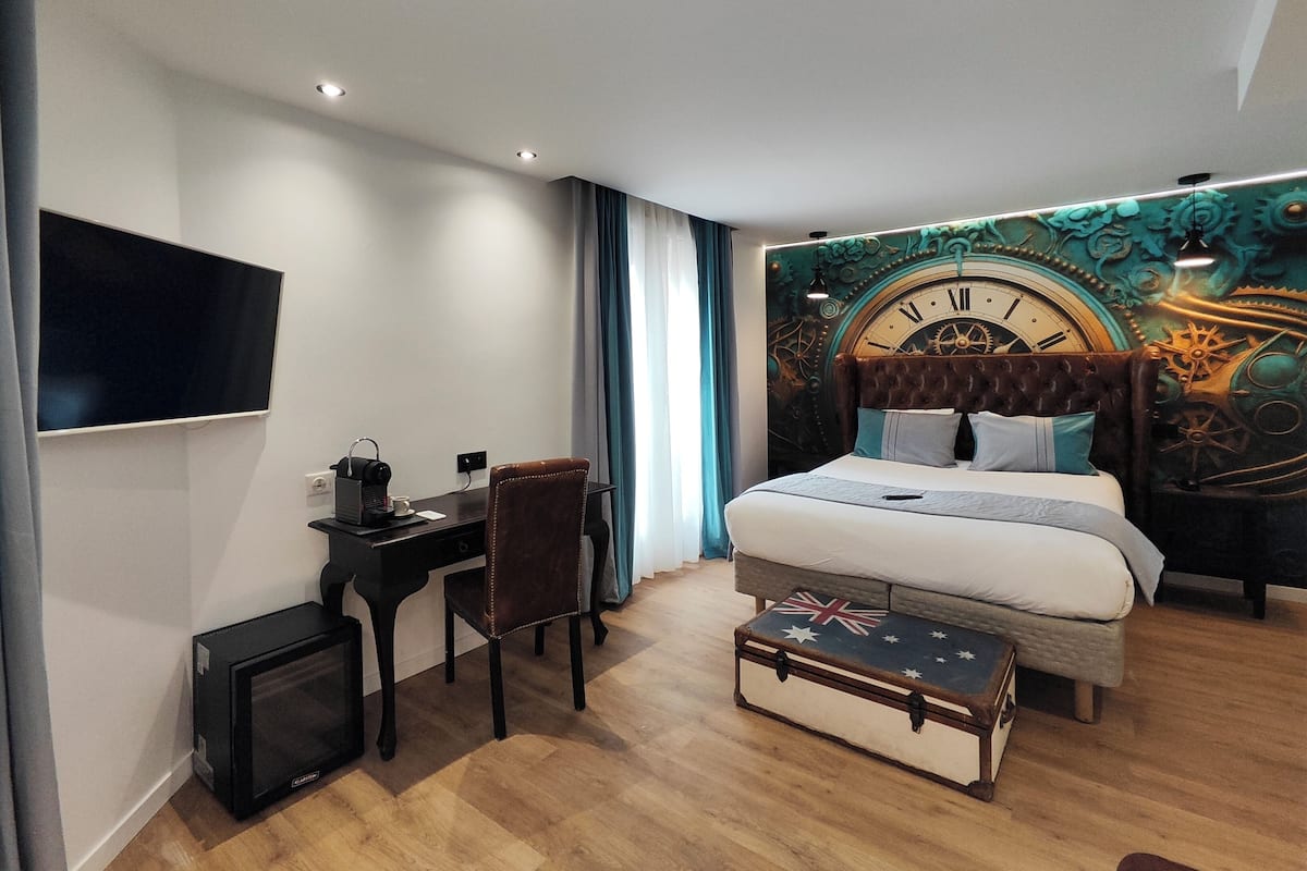 A modern hotel room with a large bed, a desk with a computer, a television, and a wall clock.