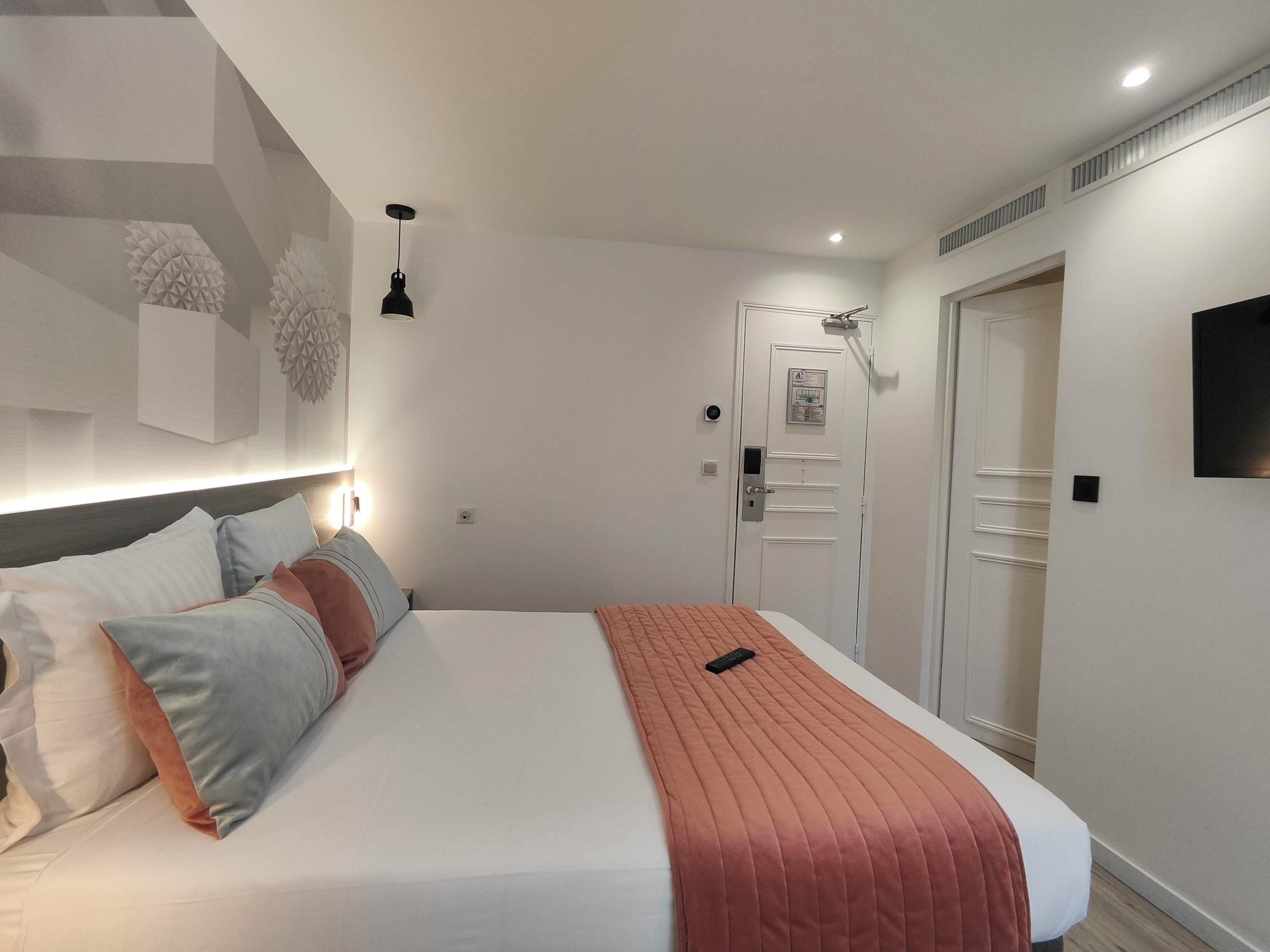Une chambre d’hôtel moderne avec un lit bien fait, une table de chevet, un téléviseur fixé au mur et une porte dotée d’un panneau de sécurité.