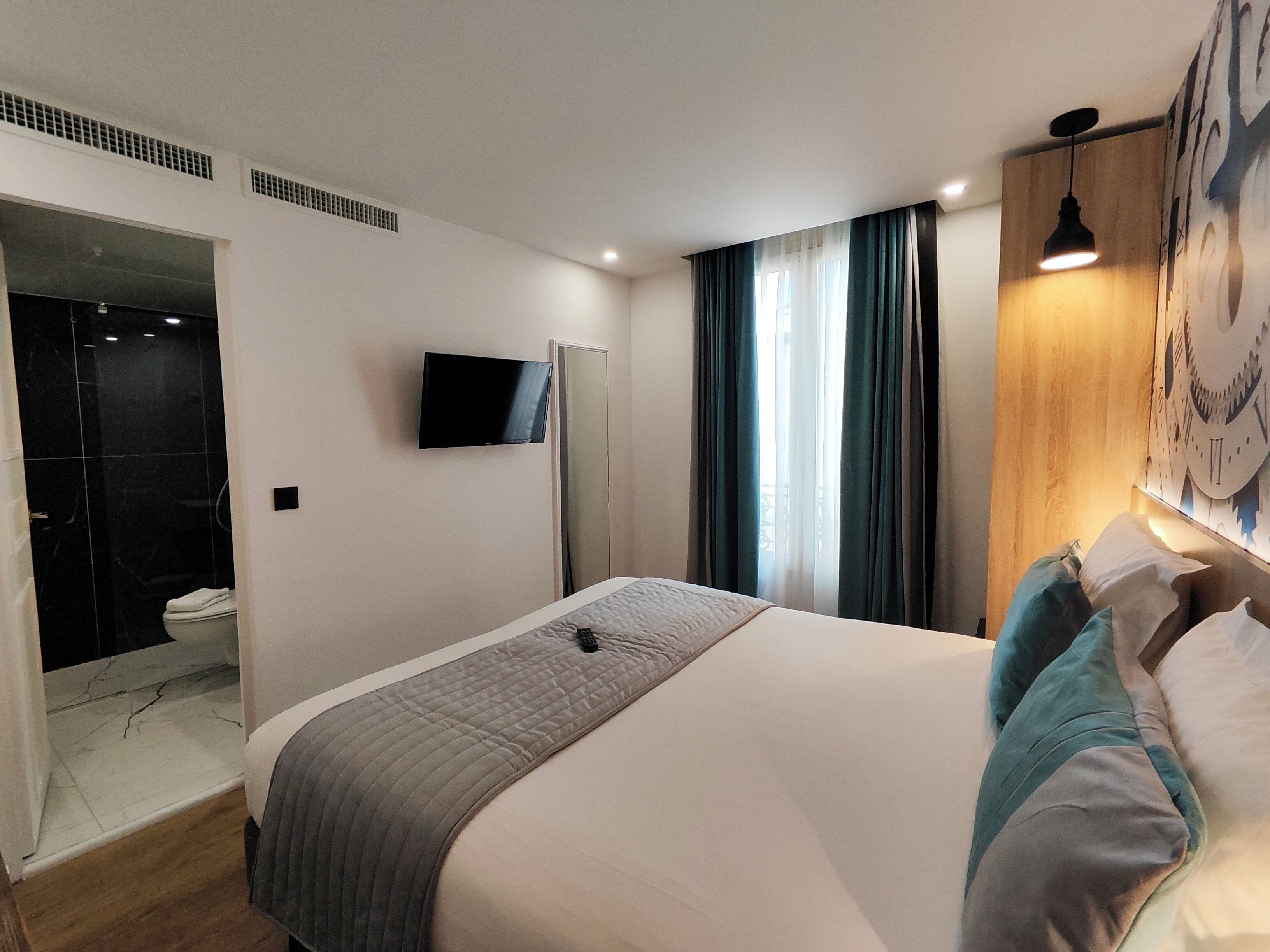 Una habitación de hotel moderna con una cama grande, un televisor de pantalla plana montado en la pared, un cabecero de madera y un ventanal con cortinas.