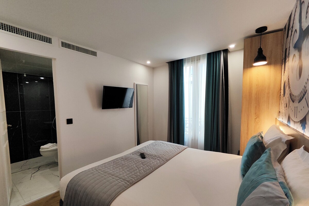 A modern hotel room with a large bed, a flat-screen TV mounted on the wall, a wooden headboard, and a large window with curtains.