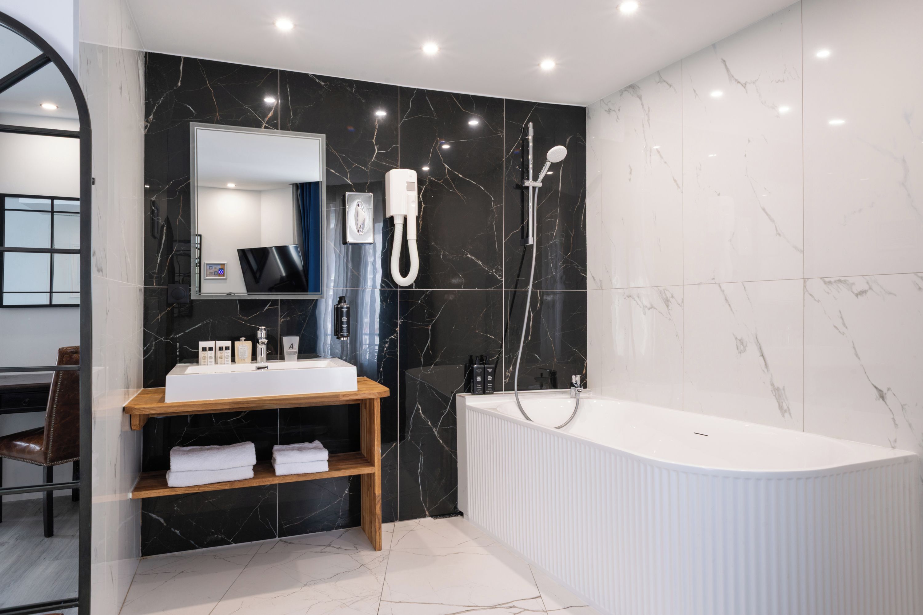 Un baño moderno con pared de mármol negro, lavabo blanco, tocador de madera, bañera grande y ducha de mano.