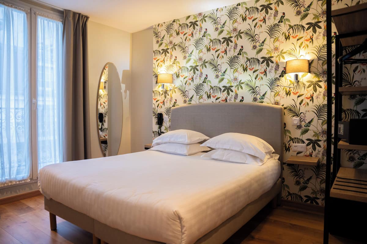 Cama bem arrumada com lençóis brancos em quarto com papel de parede floral, luminária de cabeceira e espelho na parede.