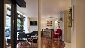 Ein modernes Wohnzimmer mit einer Couch, Sesseln, einem Couchtisch und einem roten Sessel. An der Wand ist ein Fernseher befestigt, rechts steht eine Pflanze und es gibt ein Fenster mit Vorhängen.