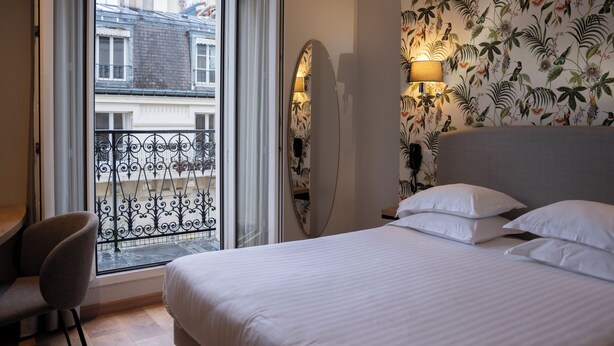 Ein Hotelzimmer mit Bett, einem Sessel, einem Balkon mit Blick auf Gebäude und einer Wand mit Blumenmuster.