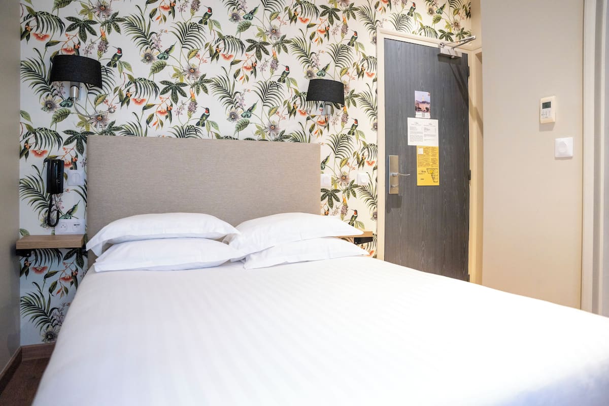Uma cama bem arrumada com lençóis brancos em um quarto com papel de parede floral, uma mesa de cabeceira de madeira, uma luminária na parede e uma porta com um mural de avisos.