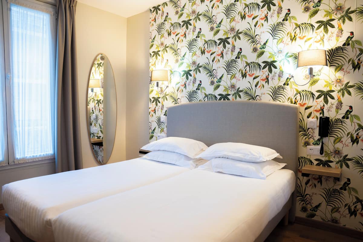 Quarto de hotel com cama de casal, um espelho grande, luminária fixada na parede e papel de parede com design tropical.