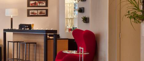 A modern bar area with a red chair, a small round table, and a bar counter. There are framed pictures on the wall and a potted plant on the right.