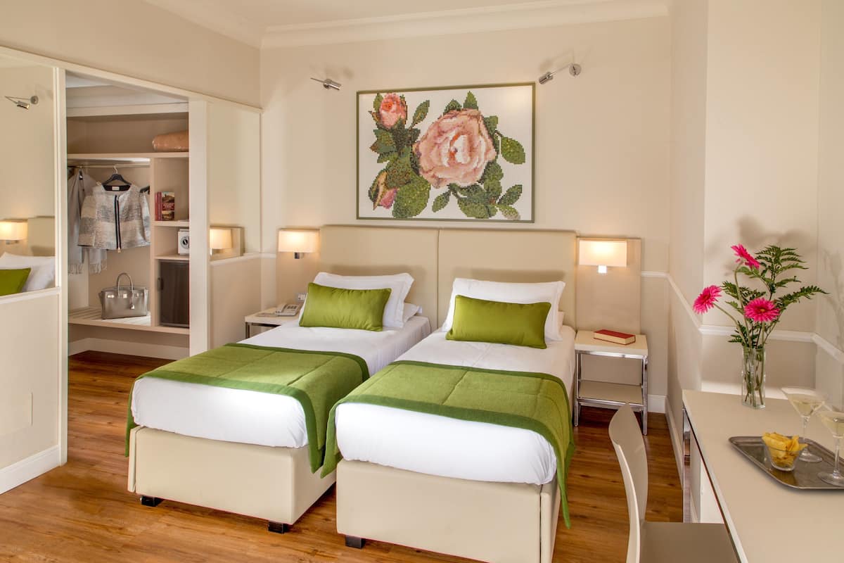 A hotel room with two beds, a desk with a tray, a vase with flowers, and a painting on the wall.