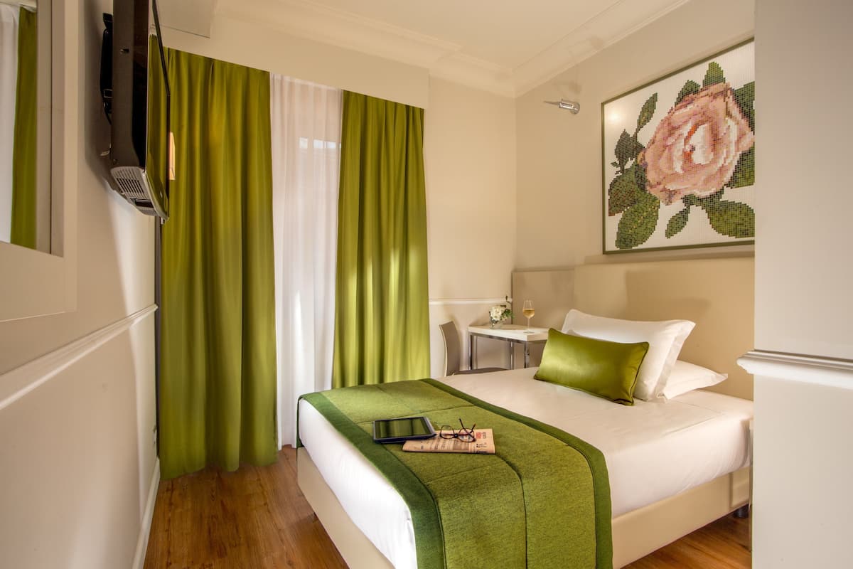 A hotel room with a bed, green curtains, a painting of a rose, a small table with a glass of wine, and a television on the wall.