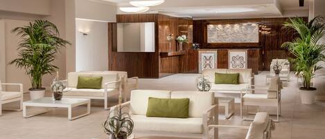 A spacious lobby with a modern design, featuring a central seating area with white sofas and armchairs, surrounded by potted plants.