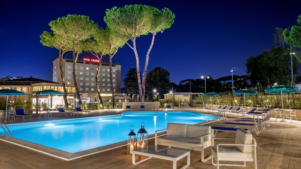 A swimming pool with lounge chairs and umbrellas, surrounded by trees and a hotel building in the background. - Hotel Cristoforo Colombo (Rome)