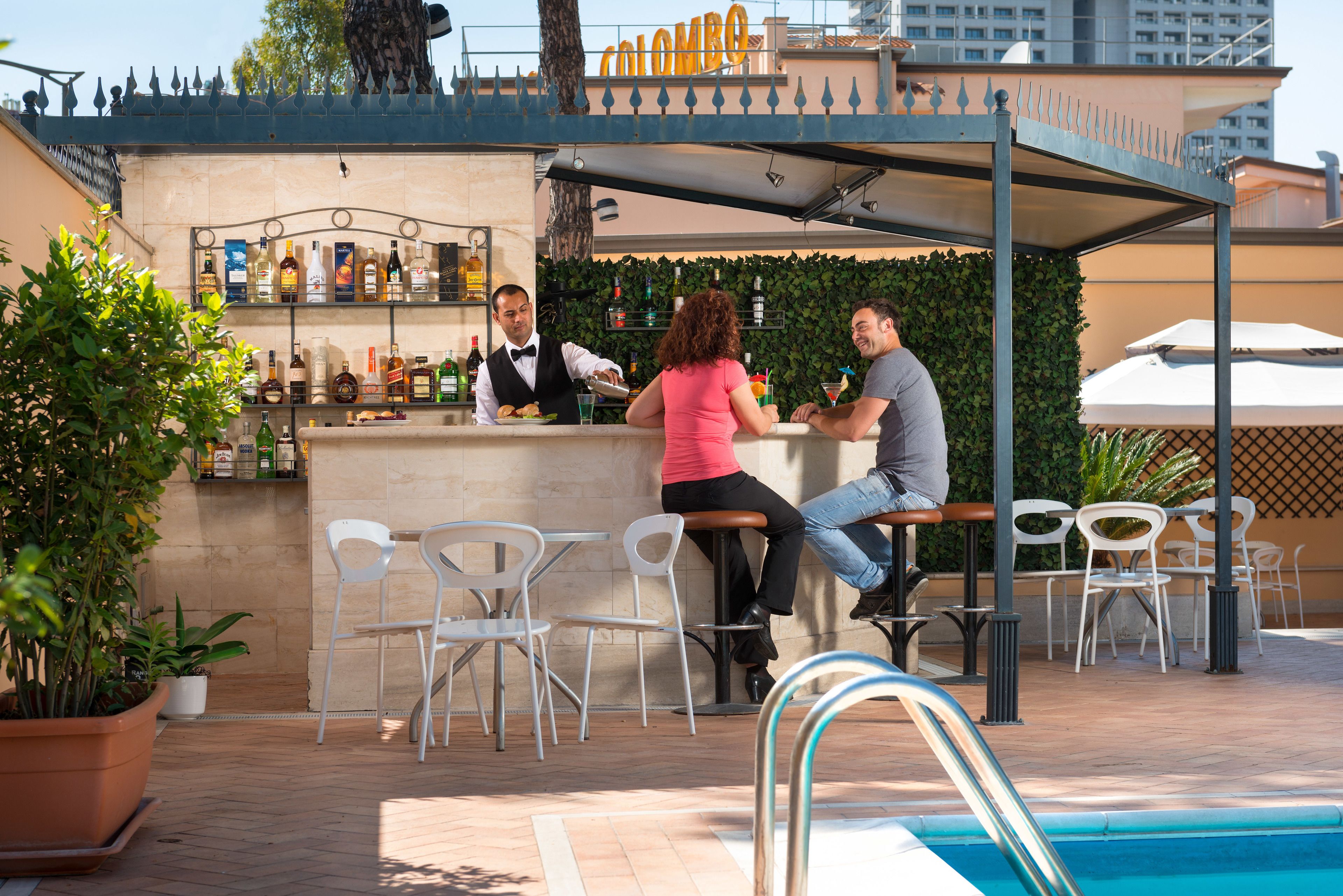 Ein Außenbereich mit Bar, Pool, Barhocker und einer Auswahl an Flaschen in Regalen.