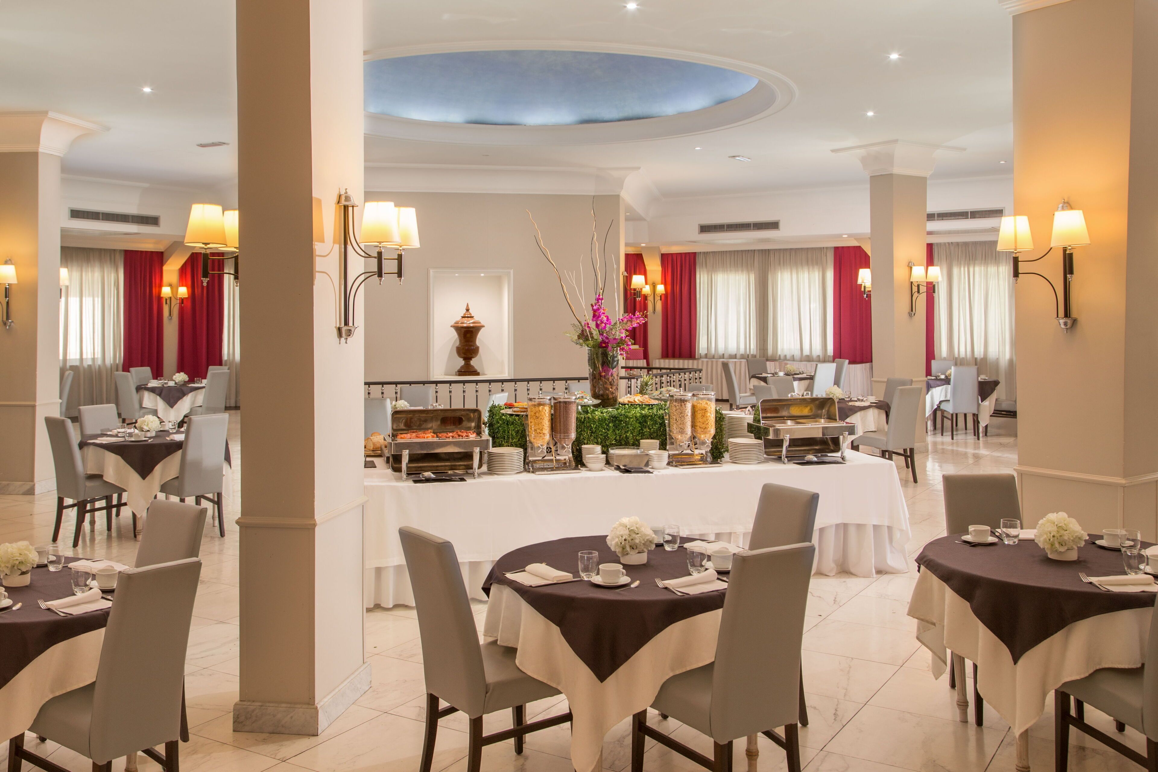 Comedor de hotel con mesas redondas, sillas y un bufé al fondo.