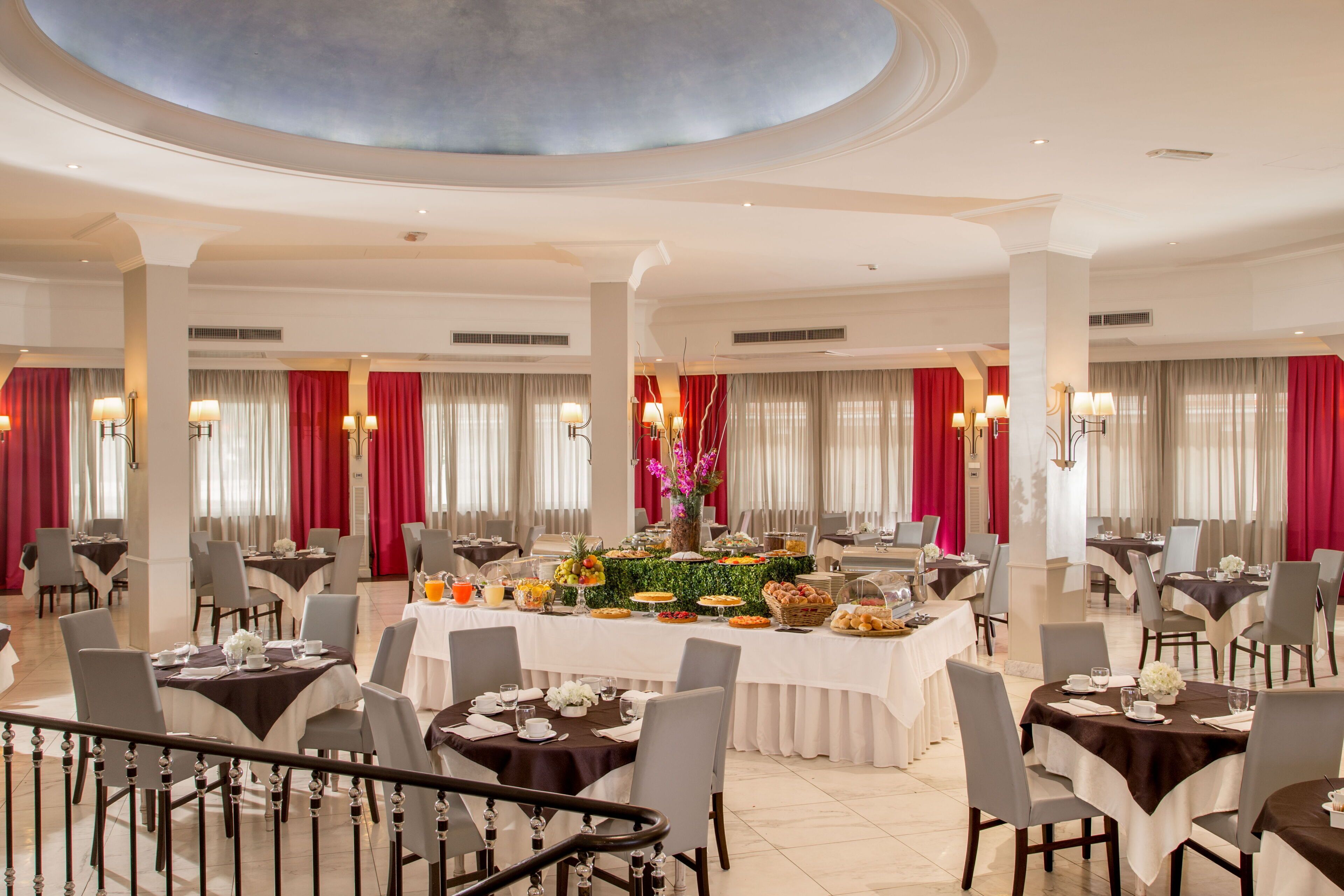A spacious dining area with round tables, red curtains, and a central buffet.