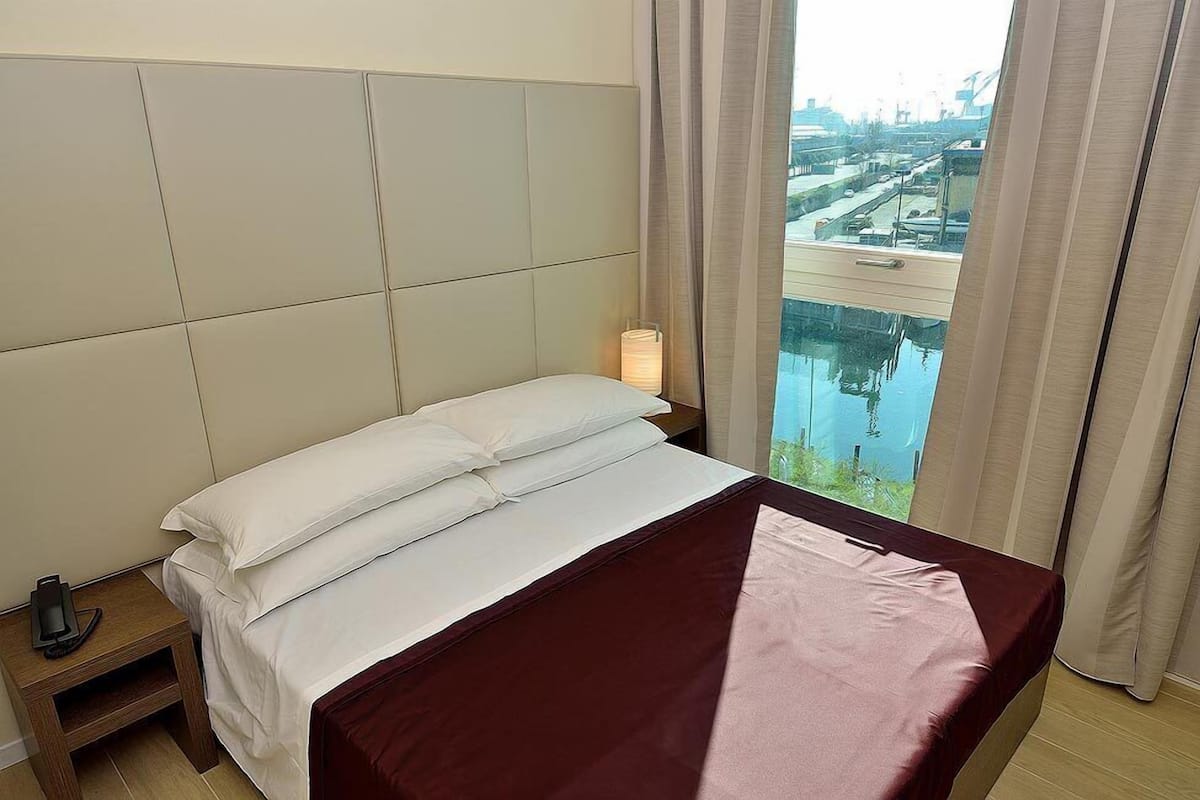 Una habitación de hotel con una cama, una mesita de noche, una lámpara y vistas a un paisaje urbano por la ventana.