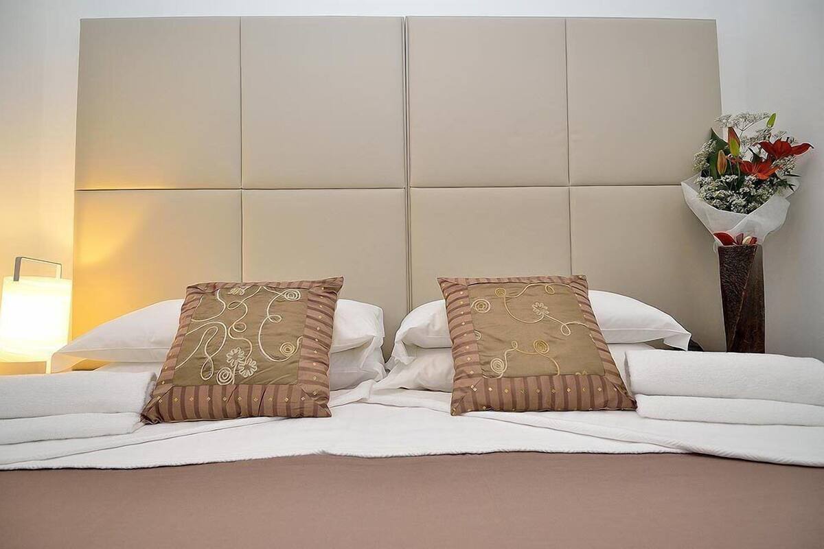 Una cama bien hecha con dos almohadones decorativos, una lámpara de noche y un florero con flores.