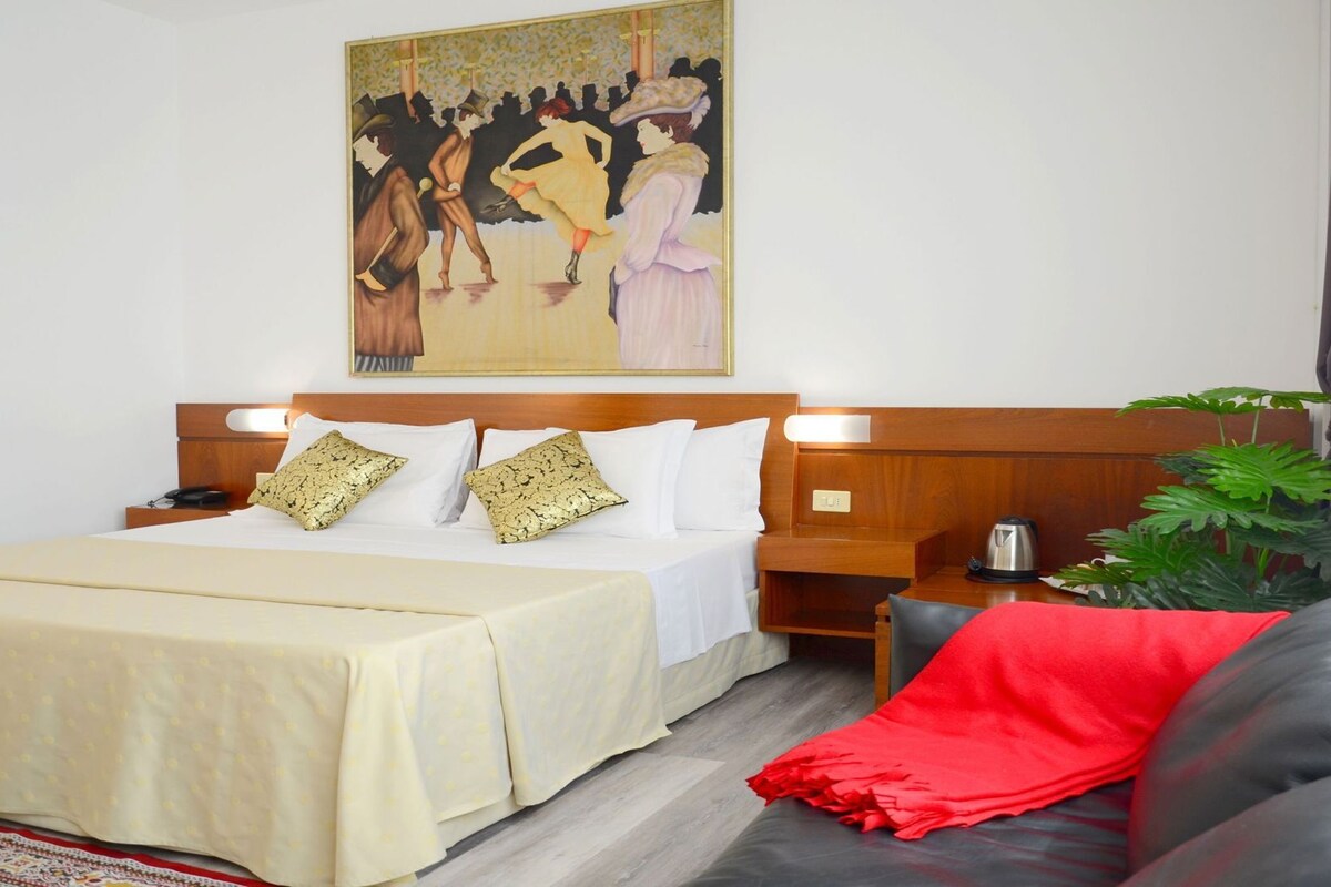 Une chambre d’hôtel avec un lit, une table de chevet, une machine à café, un plaid rouge et un grand tableau accroché au mur.