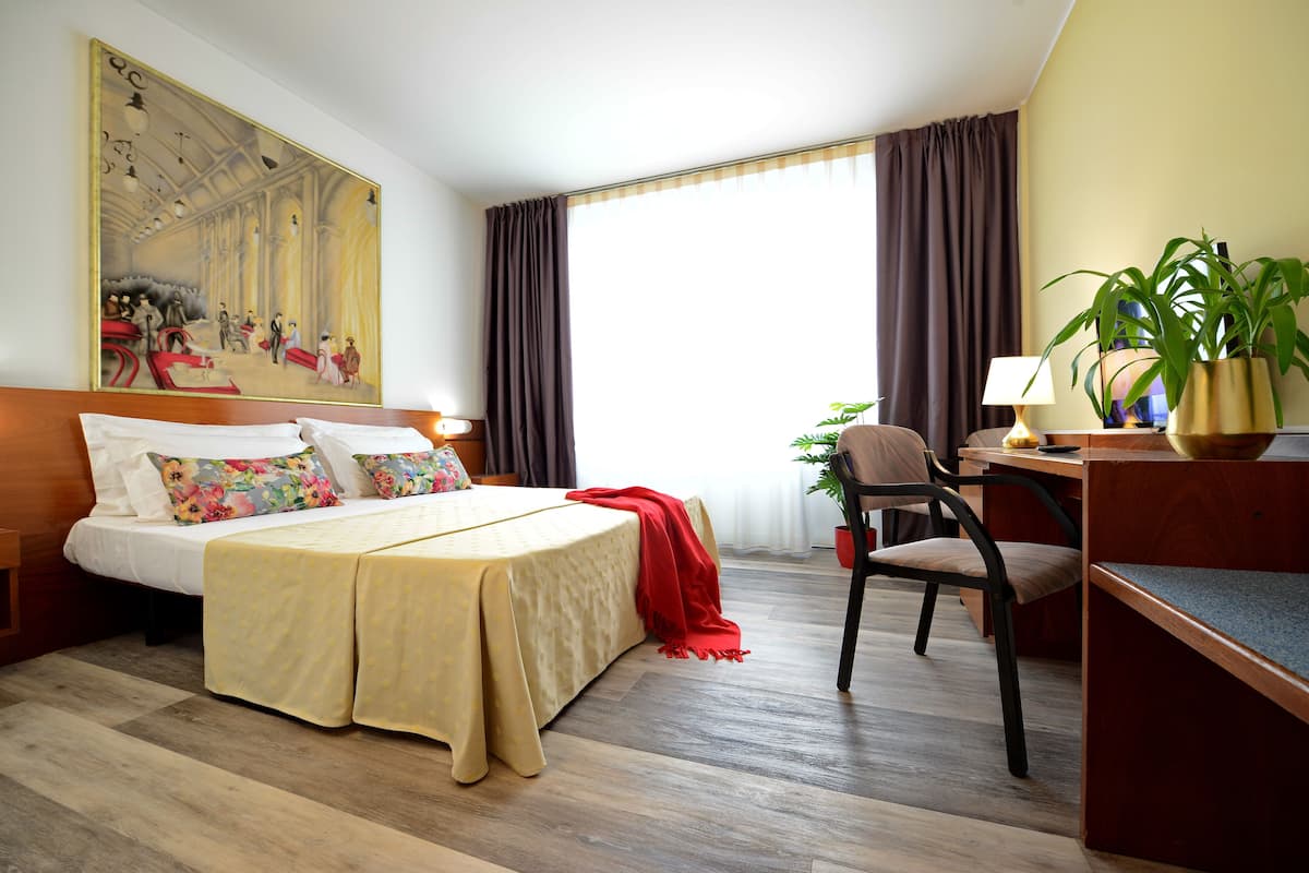 A hotel room with a bed, desk, chair, and a large painting on the wall.
