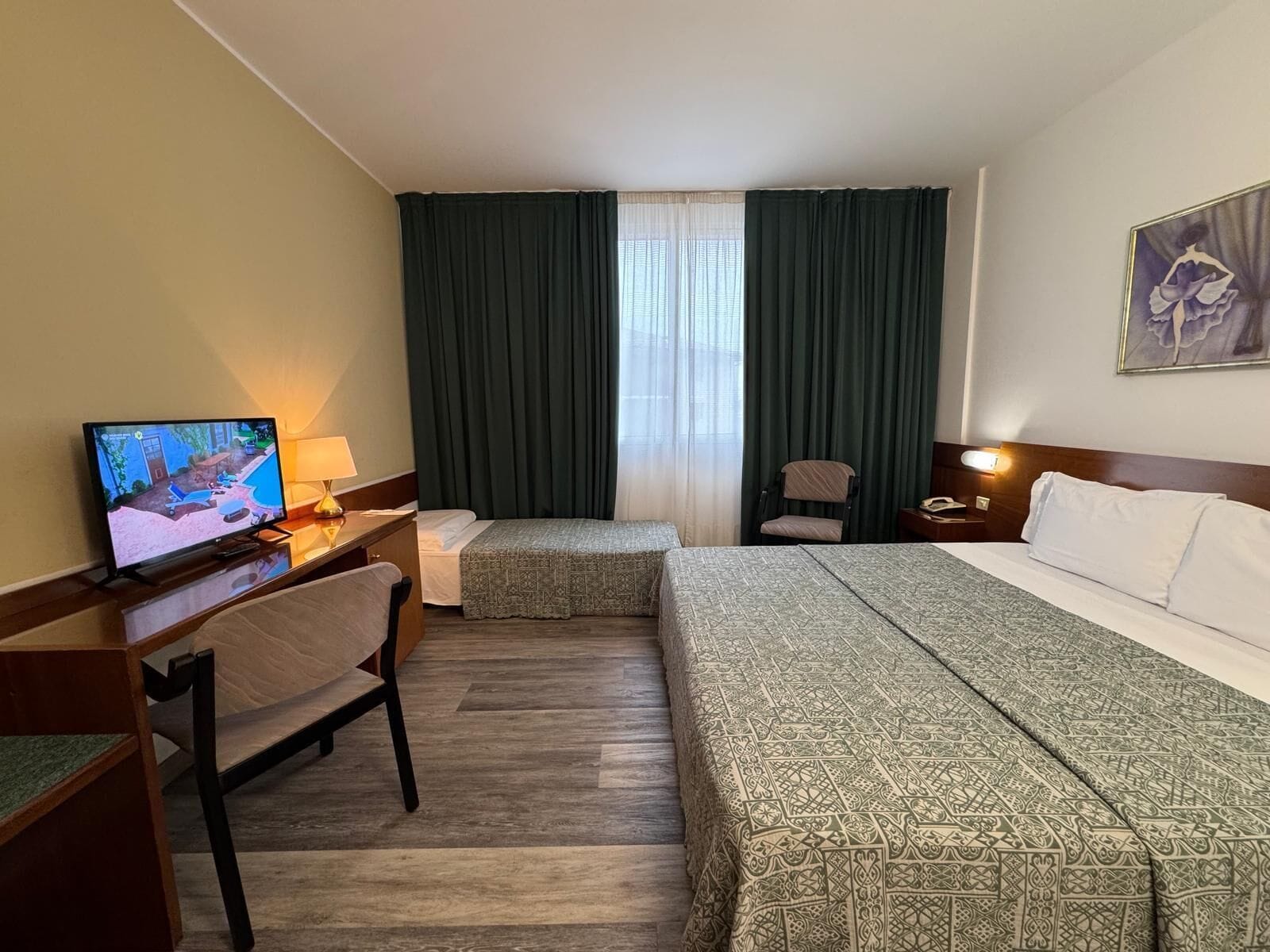 A hotel room with a bed, a desk with a TV, a chair, a bedside table, and a window with curtains.