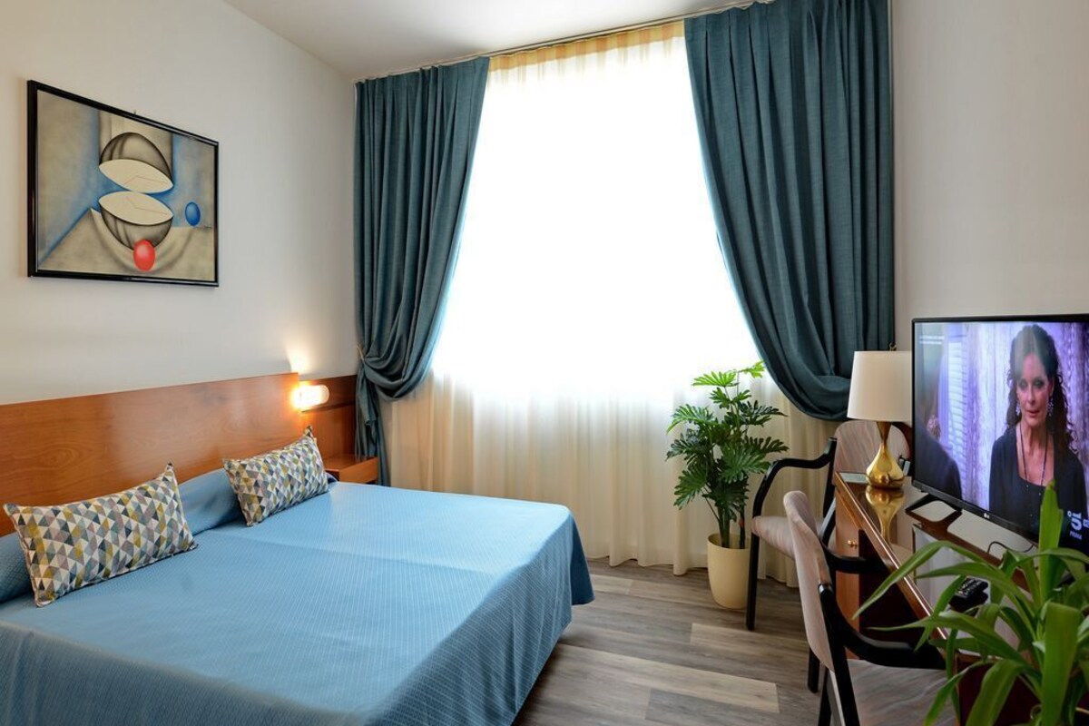 Habitación de hotel con cama, escritorio con computadora, televisor y ventana con cortinas.